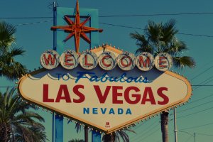 A colorful sign with a retro design reads "Welcome to Fabulous Las Vegas Nevada." The sign is topped with a red star and surrounded by palm trees, set against a clear blue sky.