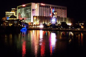 Night view of Bally’s and Flamingo hotels in Las Vegas, with colorful lights and signs reflecting on the water in the foreground. The scene is lively, showcasing illuminated buildings and a dark, peaceful sky.