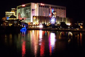 Night view of a brightly lit hotel and casino complex with colorful signage and lights reflecting on a calm body of water in the foreground. The building, adorned with large advertisements, showcases a vibrant nightlife scene. Trees and smaller structures are also visible.