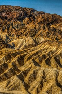 A rugged, golden-brown landscape showing eroded hills and ridges under a clear blue sky. The terrain features sharp, jagged formations with deep crevices, creating a striking texture and pattern across the scene. The distant mountain range provides a contrast in color.