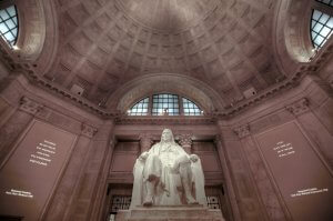 A grand statue sits within an ornate rotunda. The high ceiling features intricate detailing, lit softly by recessed lighting. The statue depicts a seated figure in historical attire, reminiscent of those commemorated for their contributions to government jobs, framed by large arched windows and text inscribed on the walls.
