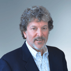 A man with curly gray hair and a beard is wearing a dark blazer over a light-colored, checked dress shirt. He is posing against a plain, light blue-gray background, exuding the calm confidence of seasoned leaders.