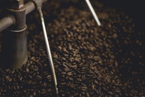 Close-up of freshly roasted coffee beans being stirred by a machine. The dark, shiny coffee beans fill the frame, while metal rods of the machine are partially visible, ensuring an even roast. The rich texture of the beans is prominently displayed.