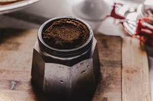 A close-up of a moka pot filled with freshly ground coffee, placed on a wooden surface. The metallic pot has a worn and rustic appearance, surrounded by a blurred background that includes a plate and what appears to be some red decorative item.