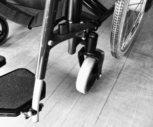 Black and white close-up photo of parts of a wheelchair, highlighting the footrest, frame, small front wheel, and large rear wheel. The wooden floor's texture is visible, emphasizing the everyday reality of living with a disability.