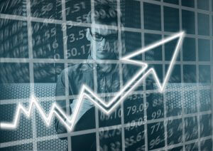 A person wearing glasses is intently looking at a tablet. The background features a large graph with numbers, stock prices, and a prominent upward arrow, symbolizing growth or success in financial markets. The image conveys themes of business analytics, economic progress, and informed decision-making.
