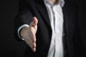 A person wearing a dark suit and white shirt is extending their hand forward, as if offering a handshake. The background is dark and out of focus, emphasizing the gesture as an invitation for a pivotal decision.