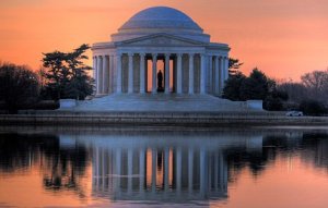 The image shows the Thomas Jefferson Memorial at sunset, with its distinctive neoclassical architecture. The dome and columns are illuminated by the orange sky, and the structure is reflected in the calm water of the Tidal Basin. Surrounded by trees and shrubs, it's a symbol adjacent to many government jobs in D.C.