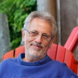 A man with gray hair, a beard, and glasses is smiling while sitting in a red Adirondack chair. Wearing a blue sweatshirt, he exudes the relaxed joy of police retirement, with lush green foliage in the background.