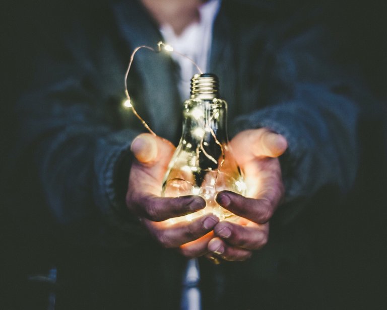 A person, like a beacon among leaders, holds a glowing light bulb containing tiny string lights with both hands. The background is dark and out of focus, making the illuminated bulb and the person's hands the focal points of the image.