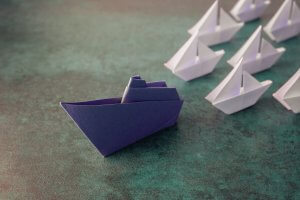 A group of small white origami boats is arranged together on a green surface. One larger purple origami boat, symbolizing leaders or leadership, is placed in front of the group, separated from the others. The scene conveys a powerful message of individuality.