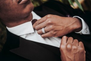 Close-up of a person dressed in a black suit adjusting their maroon tie, exuding an aura of management. The person's hands are prominently visible, showcasing a silver ring on their ring finger. The background is slightly blurred, keeping the focus on the hands and the clothing details.