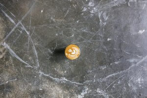 A single cup of coffee with latte art sits on a dark, scratched, and weathered surface. The latte art features a simple leaf pattern. The top-down view emphasizes the artistic coffee foam design against the rough texture of the background.