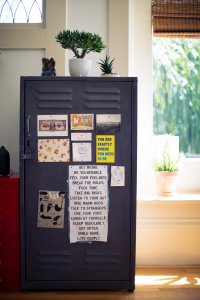 A dark gray metal cabinet adorned with various colorful magnets, postcards, and motivational quotes stands in a sunlit room, each item representing a cherished memory or goal. On top, a potted plant and small decorations are visible. The cabinet is near a window with light streaming through.