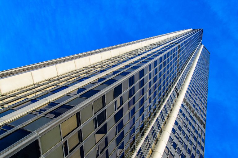 A low-angle view of a tall glass skyscraper extending into a bright blue sky. The building's facade is composed of numerous reflective windows creating a symmetrical, sleek appearance against the backdrop of a clear, cloudless daytime sky, designed to be accessible for individuals with disabilities.