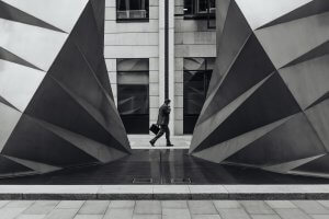 Black and white photo of a person walking between two large, angular metal structures resembling folding gates. The suited figure, carrying a briefcase, striding confidently against an urban backdrop with tall buildings and reflective surfaces, evokes the image of determined leaders in modern cities.
