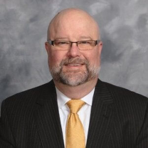 A man with glasses, a beard, and a bald head is wearing a pin-striped suit, white shirt, and a yellow tie. He is smiling and standing against a gray, cloudy background, portraying confidence as an expert in information governance.