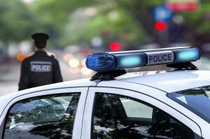 A police car with blue and white emergency lights parked on a street. In the background, a police officer stands with their back turned, dressed in a uniform and cap. The background is blurred, showing trees and indistinct street details—a serene scene perhaps marking an end to years of service and approaching police retirement.