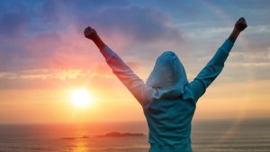 A person in a hoodie stands with arms raised triumphantly, facing a vibrant sunset over the ocean. The sky is filled with a mix of warm colors from the setting sun, casting a serene and inspirational atmosphere—an emblem of achieving one's goals.