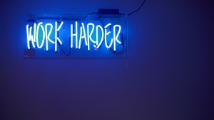 A bright, blue neon sign mounted on a wall displays the words "WORK HARDER." The letters, formed by glowing neon tubes, stand out against the dimly lit, dark blue background. The overall scene emits a motivating and energetic vibe, pushing everyone towards their goal.
