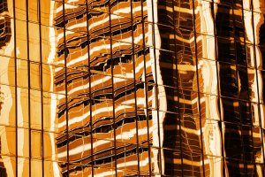A detailed close-up of the exterior of a glass building, showing a reflection of another building with a wavy and distorted pattern. The image features warm, surreal tones of gold and orange, giving it a striking and abstract visual appearance that challenges perceptions much like perspectives on disability.