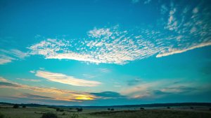 A serene landscape featuring a wide, open field with a low horizon. The sky above is mostly clear with scattered, wispy clouds illuminated by the warm colors of the setting sun, casting a soft glow over the scene.