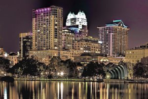 A nighttime cityscape of downtown Orlando with illuminated skyscrapers reflected in a calm lake, Orlando’s vibrant lights accent the buildings, while trees line the water and add to the colorful scene.