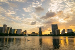 City skyline at sunset reflected on a calm lake, with two swans swimming in the foreground and a dramatic sky filled with clouds overhead.