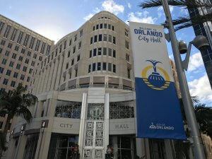 Orlando City Hall building with large windows and a decorative entrance. A blue and yellow banner on a pole reads "Welcome to Orlando City Hall" with the city logo, palm trees, and a partly cloudy sky.