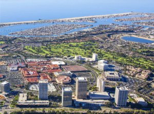 Aerial view of a coastal city with high-rise buildings, green golf courses, a shopping area, and a marina, with the ocean and boats in the background under a clear blue sky.