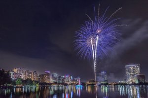 Fireworks burst in the night sky over a city skyline with illuminated buildings, reflecting on a calm lake below. The scene is vibrant and festive, with purple and white fireworks as the focal point.