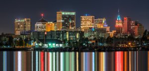 The skyline of a city at night, featuring a variety of illuminated tall buildings reflecting in the calm water below. The vibrant lights of the buildings create colorful streaks on the water, showcasing a mix of modern and historical architecture.