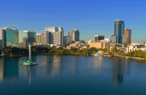A city skyline with modern high-rise buildings is reflected in a lake. A large fountain sprays water in the lake’s center, and a colorful rainbow arch sits near the waterfront under a clear blue sky.