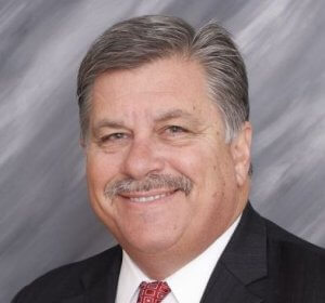 A middle-aged man with a mustache and short gray hair is wearing a dark suit, white shirt, and a red patterned tie. Smiling confidently in front of a gray, softly-textured background, he embodies the poise of someone versed in succession planning.