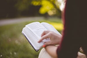An adult is sitting on the grass outside, holding an open Bible. They are pointing to a passage in the Book of Colossians. The background is green and blurred, indicating a park or garden.