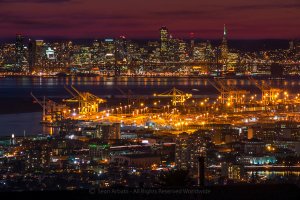 Nighttime skyline of a bustling city with illuminated buildings and cranes at a port. The scene is set against a deep purple and orange sky, reflecting off the water. Detailed structures and lights create a vibrant, urban landscape.