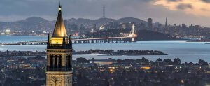 A cityscape at dusk showing a tall clock tower in the foreground, with a lit bridge stretching across water toward a city skyline and distant hills under a cloudy sky.