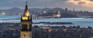 A view of the San Francisco skyline at dusk with the Bay Bridge prominently lit up, connecting the city to land across the water. In the foreground, a tall, illuminated clock tower stands above the surrounding urban landscape.