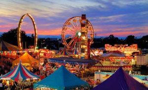 Colorful carnival tents, a lit Ferris wheel, and a looping ride are set against a vibrant sunset sky, with crowds of people enjoying the festive atmosphere.
