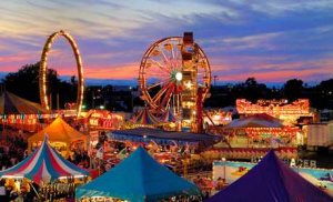 A vibrant carnival at sunset featuring colorful tents, a Ferris wheel, and various amusement rides. The glowing lights contrast against the pink, orange, and purple hues of the sky, creating a lively and festive atmosphere.