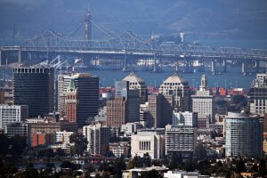 A cityscape with tall downtown buildings in front, a large bridge spanning water in the background, and hazy mountains beyond.