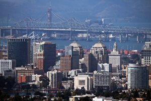 Aerial view of downtown Oakland, California, featuring a mix of modern and historic buildings. The San Francisco–Oakland Bay Bridge is visible in the background spanning the bay with mountains in the distance.