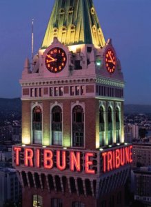 The image shows a tall clock tower at night, illuminated by lights. The tower features two large clocks and a sign with the word "TRIBUNE" in red, neon letters. The background reveals a cityscape with faintly visible buildings and a dark sky.