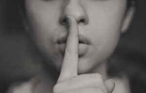 Close-up black-and-white photo of a person holding their index finger to their lips, signifying silence or a request to be quiet. The background is largely out of focus, emphasizing the person's gesture, as if reminding the viewer of the quiet needed during an interview.