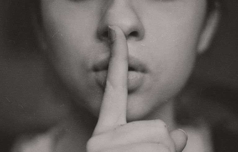 Close-up black-and-white photo of a person holding their index finger to their lips, signifying silence or a request to be quiet. The background is largely out of focus, emphasizing the person's gesture, as if reminding the viewer of the quiet needed during an interview.