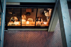 Aerial view through the window of three people sitting inside a coffee shop. They are seated at a wooden bar, focusing on reading and writing. The cozy and safe atmosphere contrasts with the brick street visible outside, framed by concrete structures.