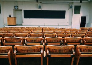 An empty lecture hall with rows of wooden seats facing a large chalkboard at the front. A wooden podium is positioned to the left, with speakers and lights mounted on the walls. The room, designed for adult learners, features a white and beige color scheme.