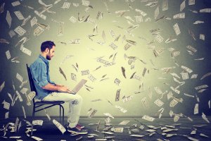 A man sits on a chair typing on a laptop while surrounded by falling banknotes, enjoying the fringe benefits of making money online. The background is a plain, neutral-colored wall, and the floor is wooden.