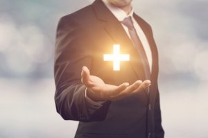 A man in a business suit holds out his hand with a glowing plus sign floating above his palm, symbolizing fringe benefits. The background is blurred, and the scene conveys a sense of positivity or adding value.
