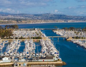Aerial view of a marina filled with numerous white sailboats docked in organized rows. The marina is surrounded by calm blue water and a distant hilly coastline under a partly cloudy sky. In the foreground, there's a dock with buildings and trees scattered around.